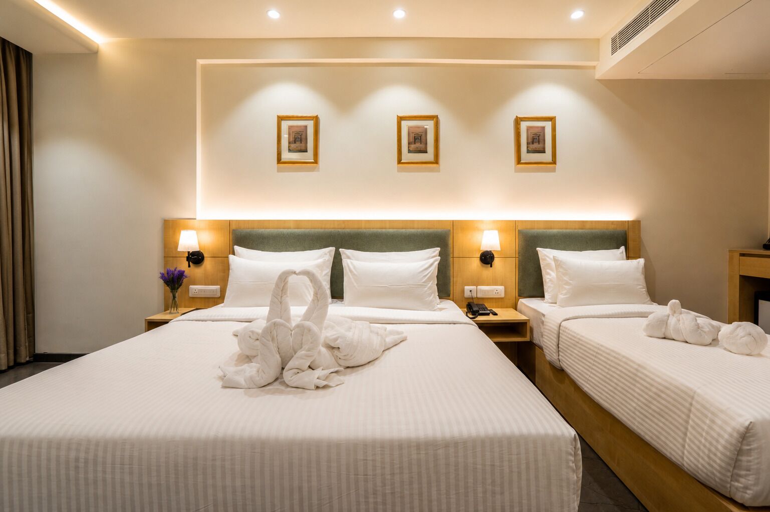 A hotel room with two beds, a headboard with wall-mounted lamps, and a wall with three framed pictures.