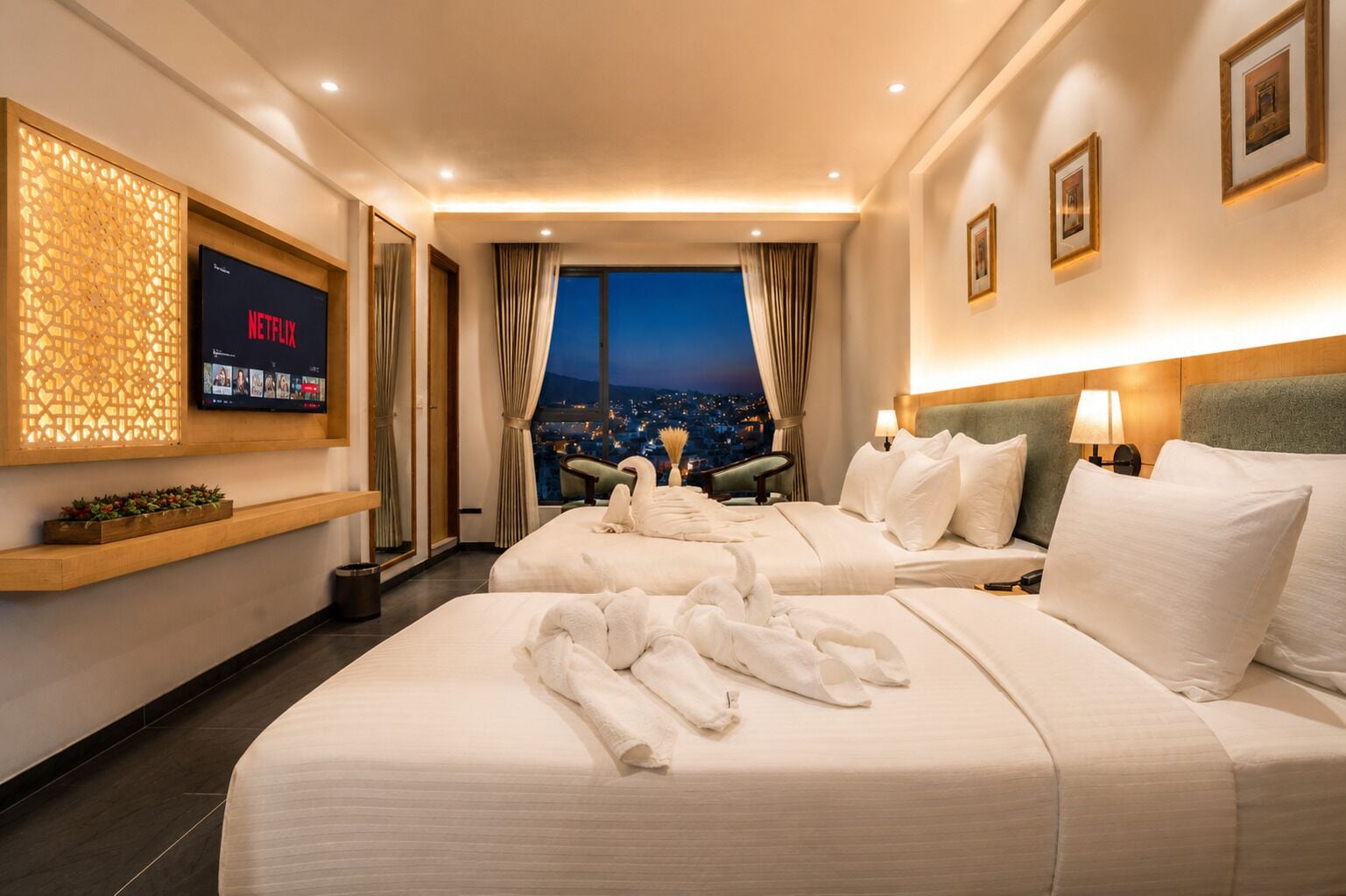 Quarto de hotel com duas camas, televisão, vista para a cidade e uma prateleira de madeira com objetos decorativos.