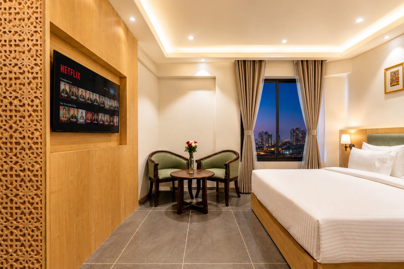 A hotel room with a bed, a desk with two chairs, a wall-mounted TV displaying Netflix content, and a cityscape view through a window.