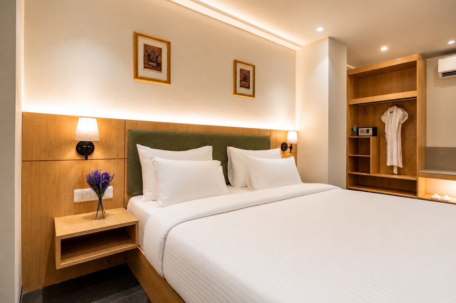Habitación de hotel con cama, lámparas de noche, mesita de noche, un jarrón con flores y un armario de madera con una bata en su interior.