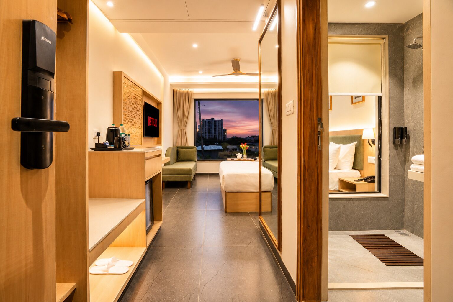 A hotel room with a bed, a desk, a sofa, a TV, and a view of the cityscape at dusk.