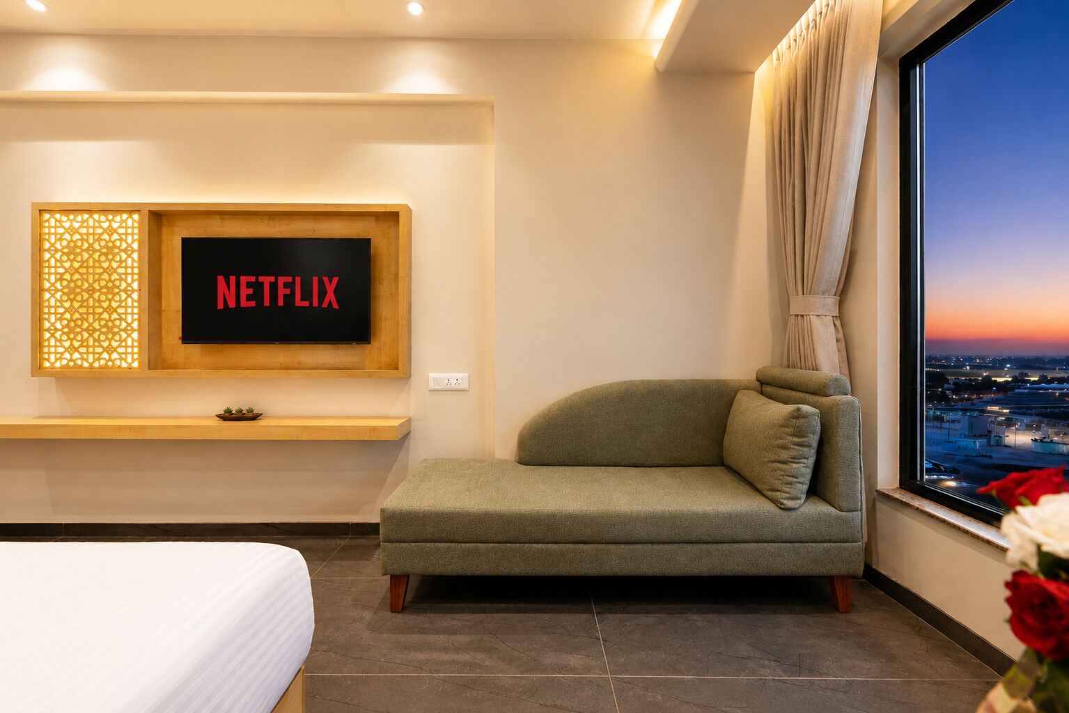 A modern hotel room with a large window, a sofa, a bed, and a wall-mounted TV displaying 'NETFLIX'.