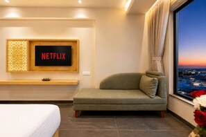 A modern hotel room with a large window, a sofa, a bed, and a wall-mounted TV displaying 'NETFLIX'. - The Avenor - Luxury Hotel in Jaipur (Jaipur)