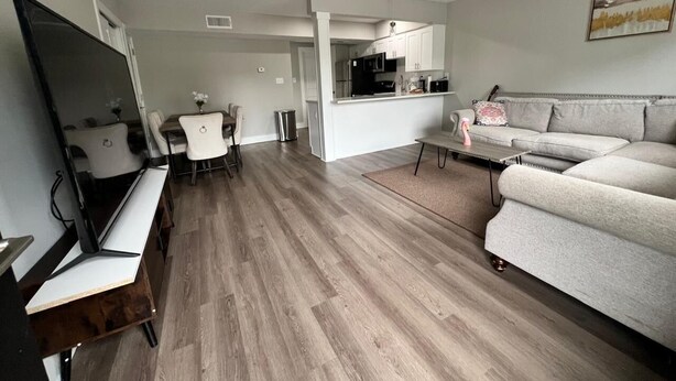 A modern living room with a flat-screen TV, a grey sofa, a dining area with white chairs, and a kitchen area with white cabinets and appliances. - Entire home ,Clean, quiet, and cozy—everything you need for the perfect stay (Virginia Beach)