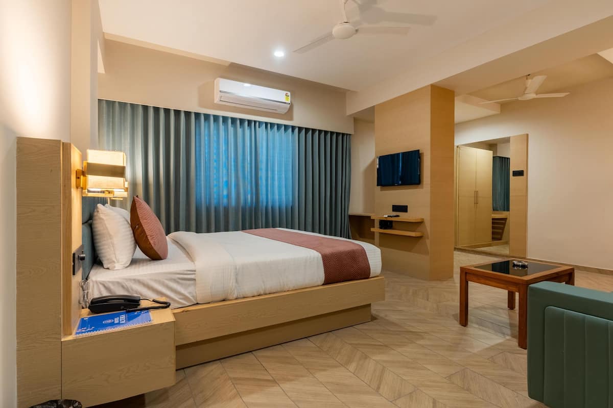 A hotel room with a bed, bedside table, lamp, air conditioning unit, TV, and a small table.