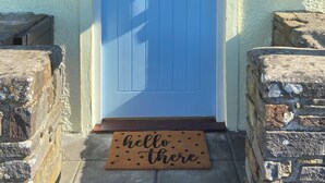 Eine blaue Tür mit kleinem Fenster, einer Fußmatte mit der Aufschrift 'Hello there', einem Briefkasten und einem Schild, auf dem 'Cowslip Cottage' steht.