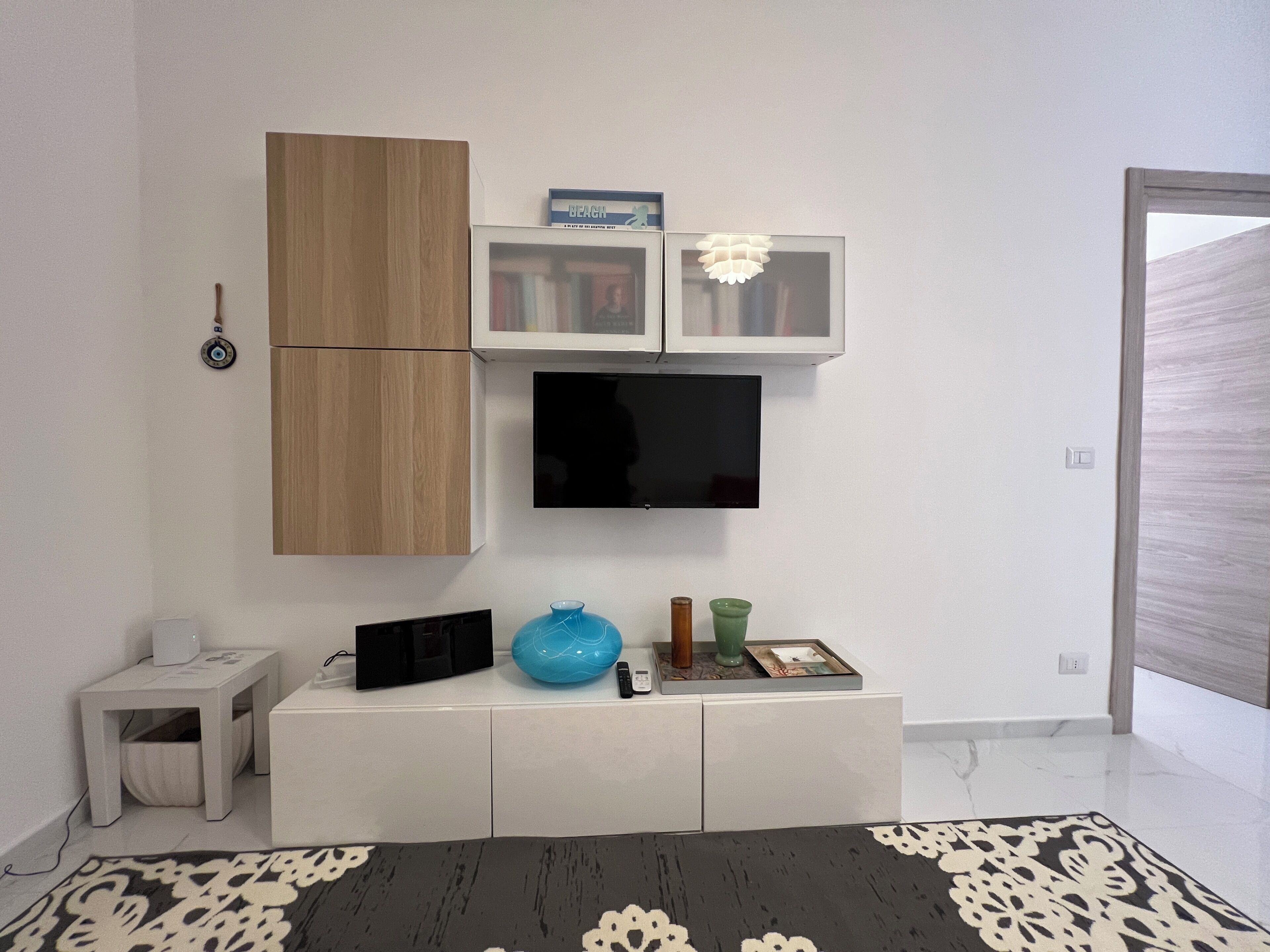 A modern living room with a flat-screen TV mounted on the wall, a wooden cabinet, a small side table, and a decorative vase.