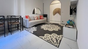 Un salon moderne comprenant un canapé, des fauteuils, une table à manger, un tapis décoratif, un ventilateur fixé au mur et un luminaire suspendu au plafond.