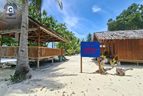 A beachside wooden structure with a thatched roof, a sign indicating a 'Sustainable Homestay', and a palm tree.