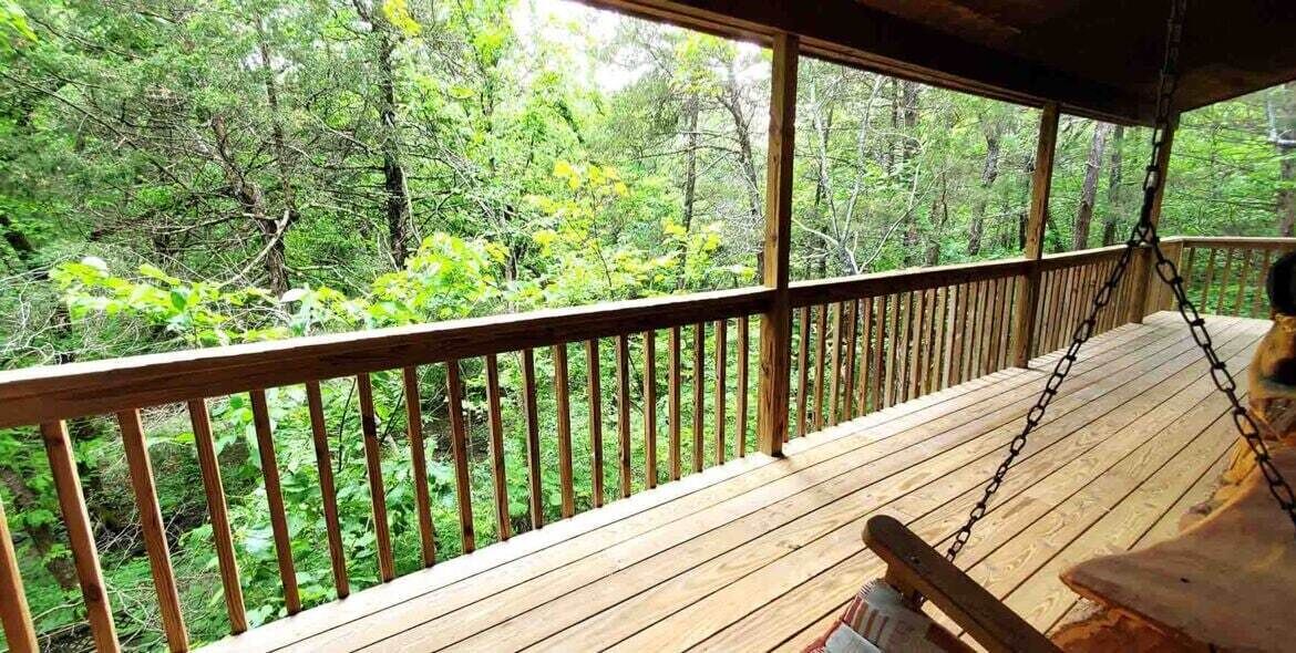 A wooden deck with a swing chair, surrounded by a wooden railing, overlooking a forested area.