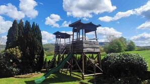 Una struttura giochi in legno con uno scivolo, in un’area erbosa con alberi e colline sullo sfondo.