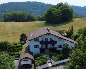A two-story house with a sloped roof, surrounded by greenery and hills.