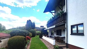 Un edificio bianco con un balcone, un piccolo giardino e una struttura con tetto rosso sullo sfondo.