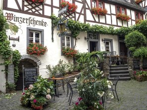 Ein traditionelles Fachwerkhäuschen mit einem Schild, auf dem 'Winzerchenke' steht, Außenplätzen und einer Steintreppe, die zum Eingang führt.