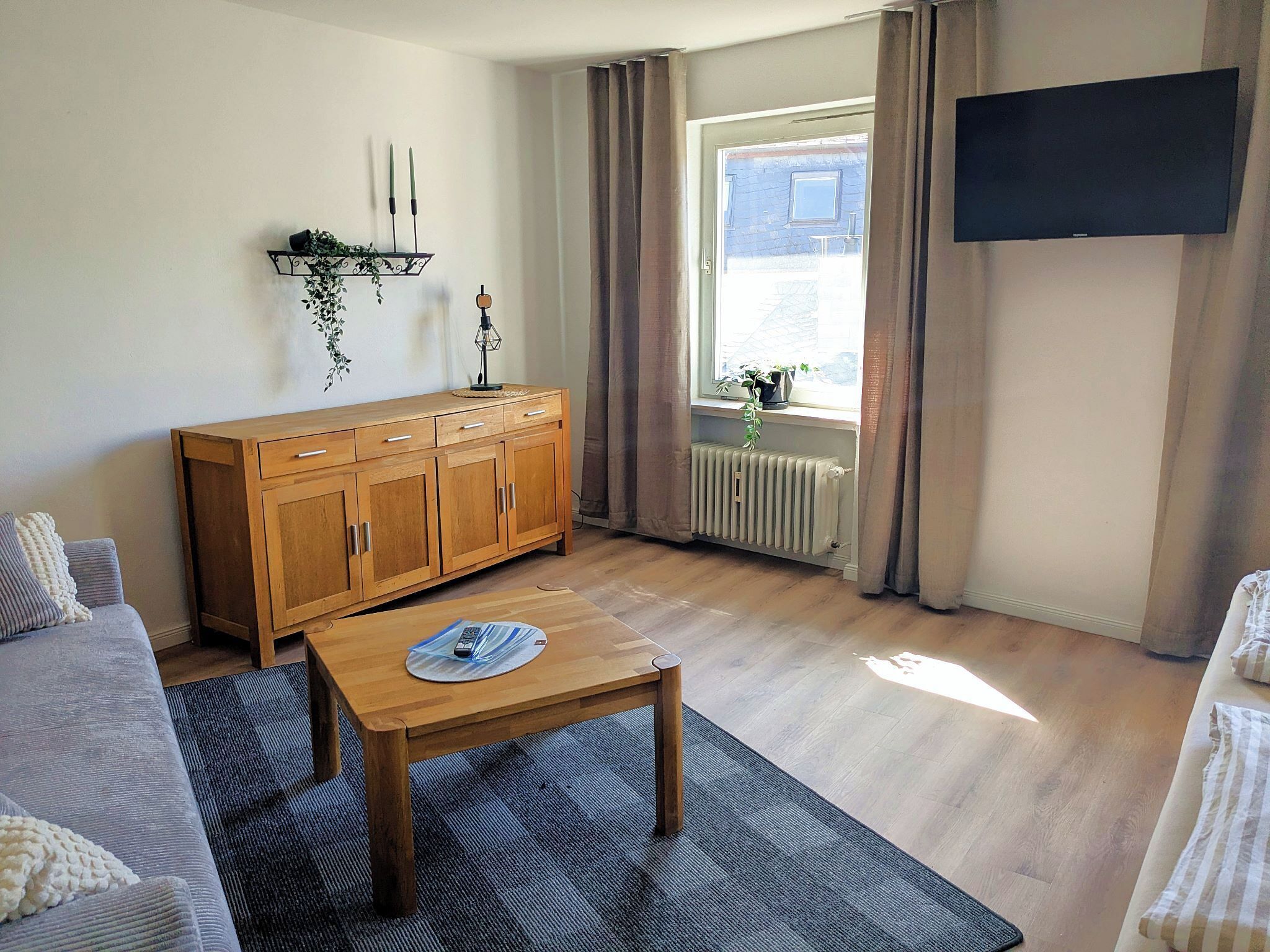 A living room with a wooden cabinet, a coffee table, a sofa, a TV mounted on the wall, and a window with curtains.