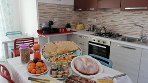 A modern kitchen with a dining table set for a meal, featuring bread, fruits, cheese, and a cake. - B&b Eden Ogliastra  (Tortolì)