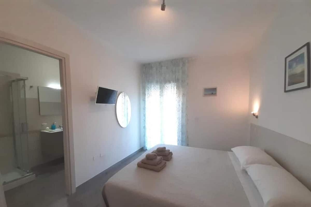 Une chambre à coucher avec un lit, une télévision, une fenêtre avec des rideaux et une salle de bain visible à travers une porte ouverte.