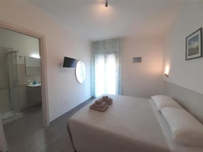 A bedroom with a bed, a TV, a window with curtains, and a bathroom visible through an open door. - B&b Eden Ogliastra  (Tortolì)