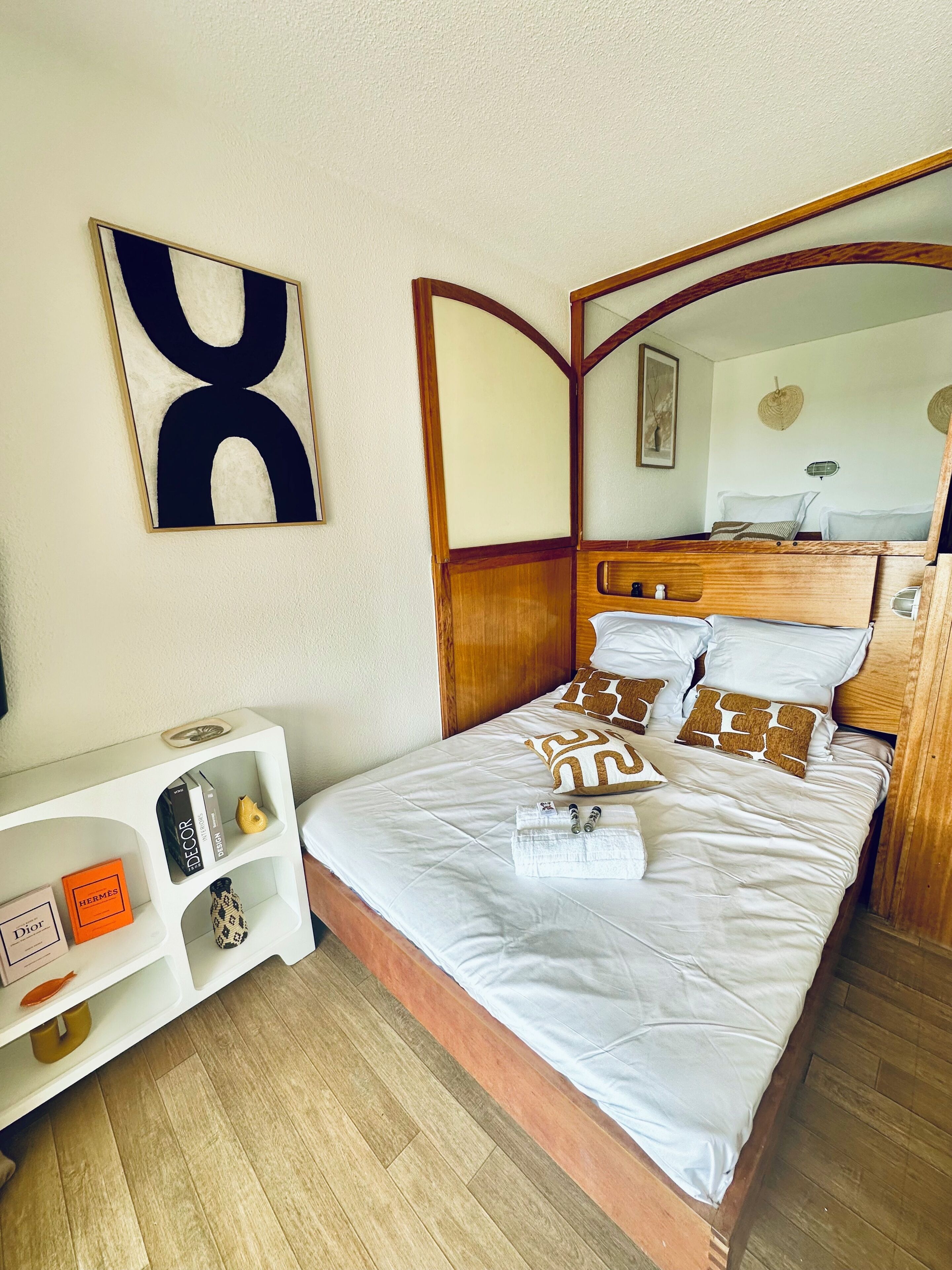 A neatly made bed with white linens and patterned pillows, a wooden headboard, a white console table with books and a vase, and a large abstract painting on the wall.