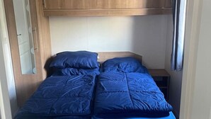 Kompakti makuuhuone matkailuautossa, jossa on kerrossänky, yläpuolella puiset kaapit ja sängyn vieressä pieni pöytä.