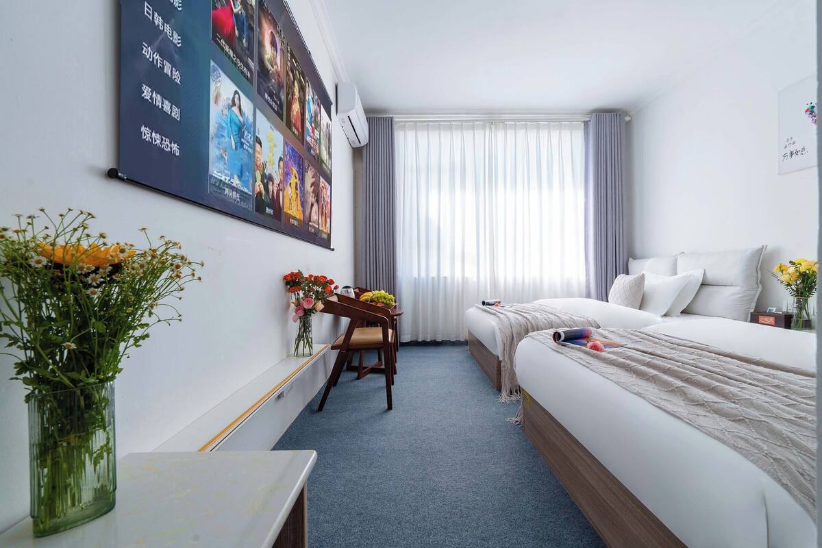 Ein Hotelzimmer mit Bett, Schreibtisch mit Stuhl, Blumen in einer Vase und einem großen Poster an der Wand.
