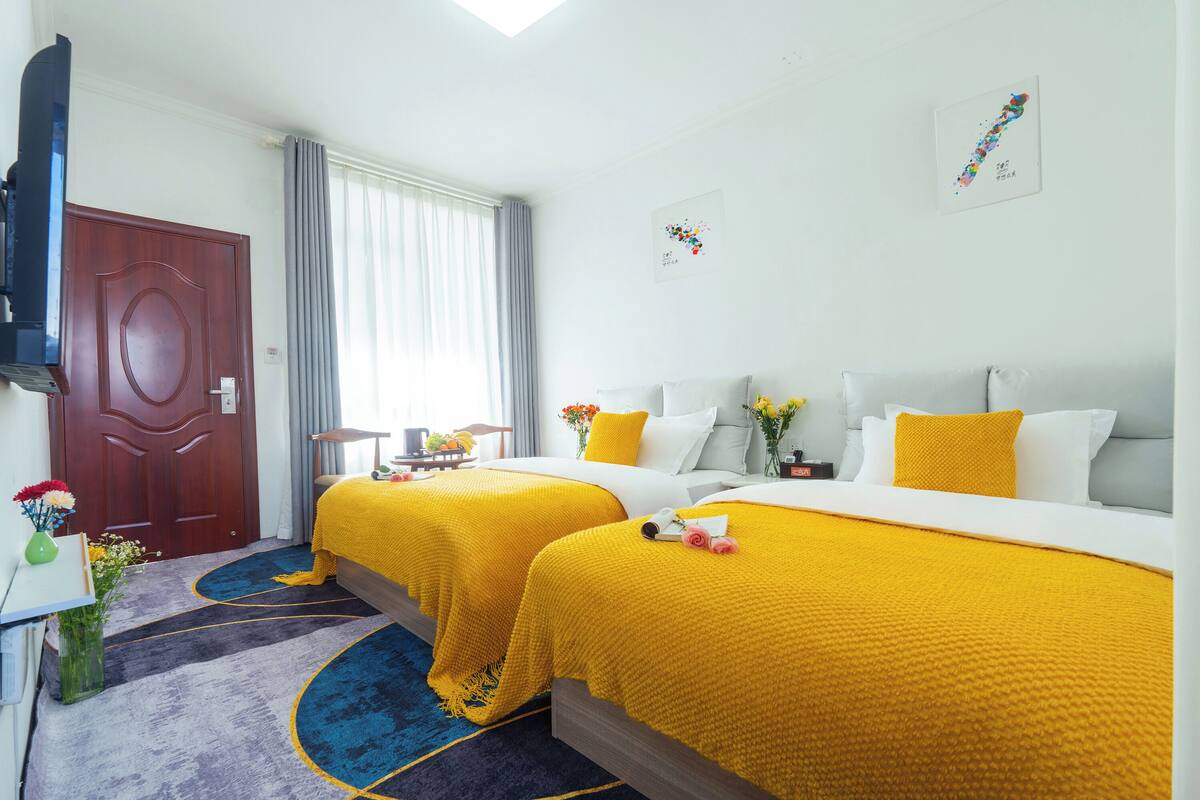 Ein Hotelzimmer mit zwei Betten, einem Fernseher, einem Schreibtisch mit Blumenvase und einem Fenster mit Vorhängen.