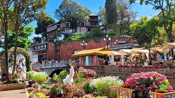 A courtyard with a wooden planter filled with various plants, a traditional building with a tiled roof, and a stone bridge in the background. - 166 Hotel-Ancient City Mufu Branch (Lijiang)