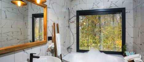 Un baño moderno con pared de mármol, una bañera con burbujas, un tocador de madera, un espejo grande y una ventana con vistas a árboles.