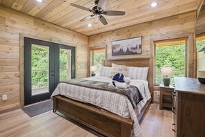 Ein Schlafzimmer in einer Holzhütte mit Bett, Deckenventilator, Nachttisch und Fenster mit Blick auf Grün.