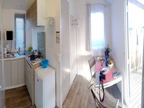 A compact kitchen with a sink, stove, and refrigerator, a dining area with a table and chairs, and a window with curtains. - Cozy mobile home - 2 guests (Sarzeau)
