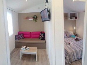A small, modern room with a sofa, a small table, a bed with a striped bedspread, and a mounted TV. - Cozy mobile home - 2 guests (Sarzeau)