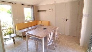 A modern kitchen with a wooden table, white chairs, a built-in bench, and a window with blinds. - Mobile home, pets welcome - 4 guests (Chaillé-les-Marais)