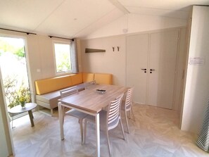 A modern kitchen with a wooden table, white chairs, a built-in bench, and a window with blinds. - Mobile home, pets welcome - 4 guests (Chaillé-les-Marais)