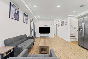 Una sala de estar moderna con un sofá gris, una mesa de centro, un televisor de pantalla plana y una zona de cocina con refrigerador.