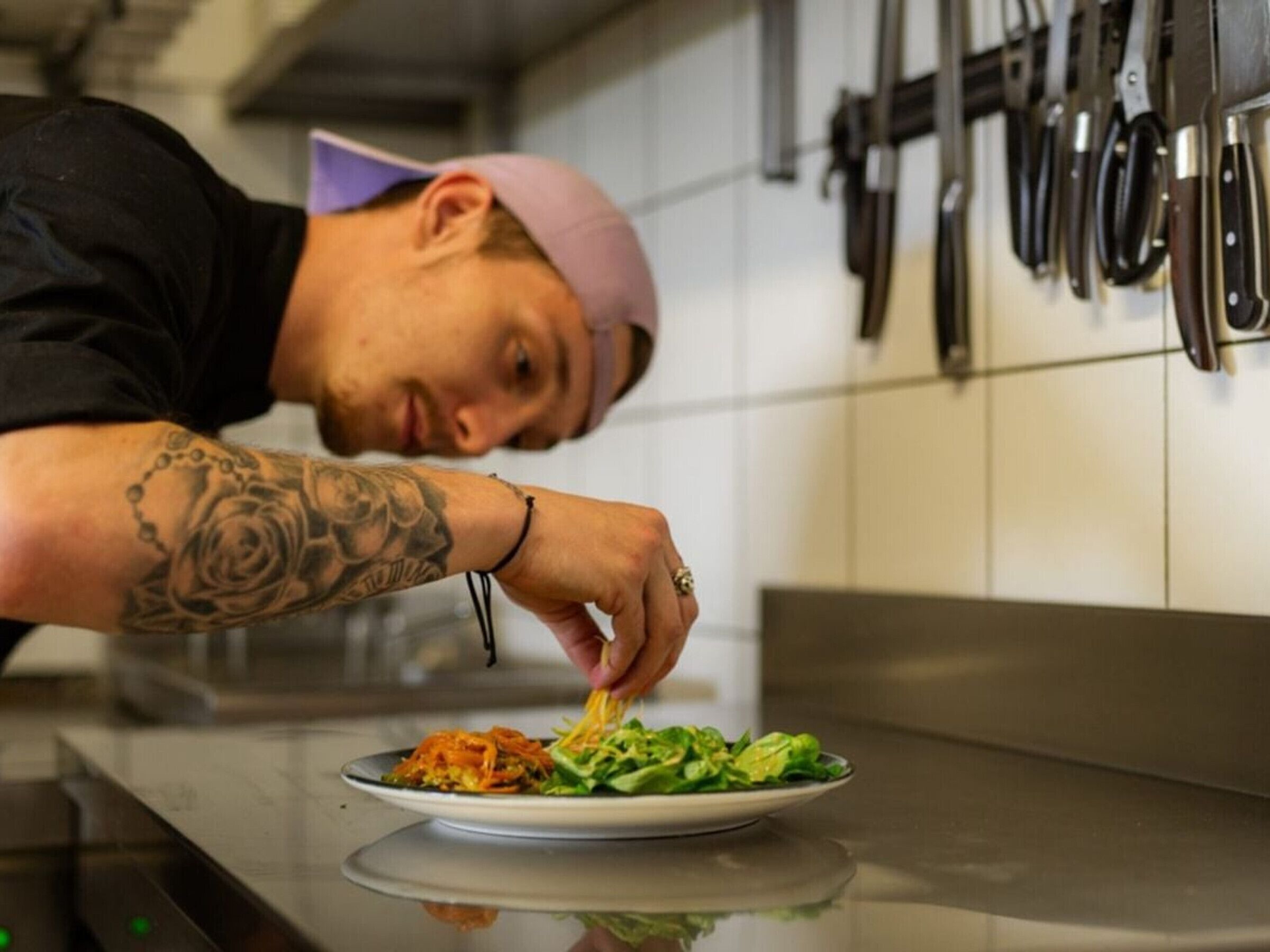 En person i ett kök, med en tallrik mat framför sig, en tatuering på armen och köksredskap upphängda på väggen.