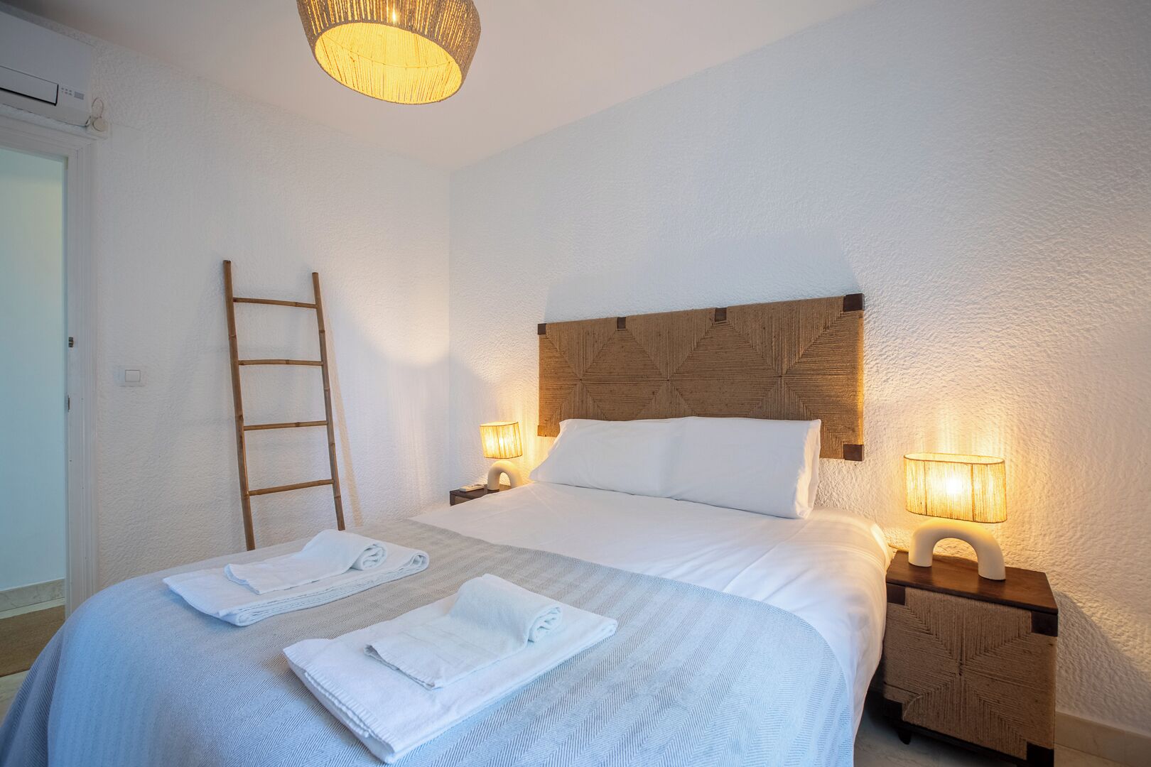 Une chambre à coucher avec un lit, deux lampes de chevet, une tête de lit en bois, une échelle et deux serviettes pliées.