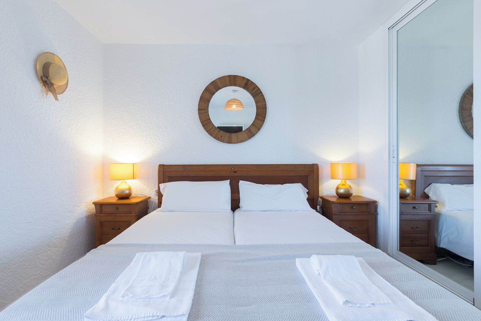 A hotel room with a large bed, two bedside tables with lamps, a round mirror on the wall, and a large mirror on the door.