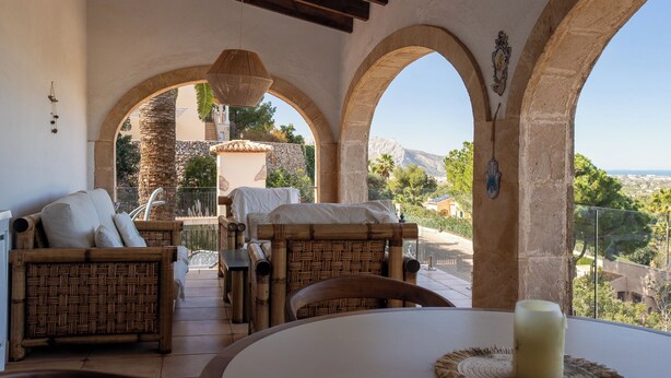 Un patio iluminado por el sol con mobiliario de mimbre, una mesa con una vela y vistas a un paisaje montañoso a través de arcos.