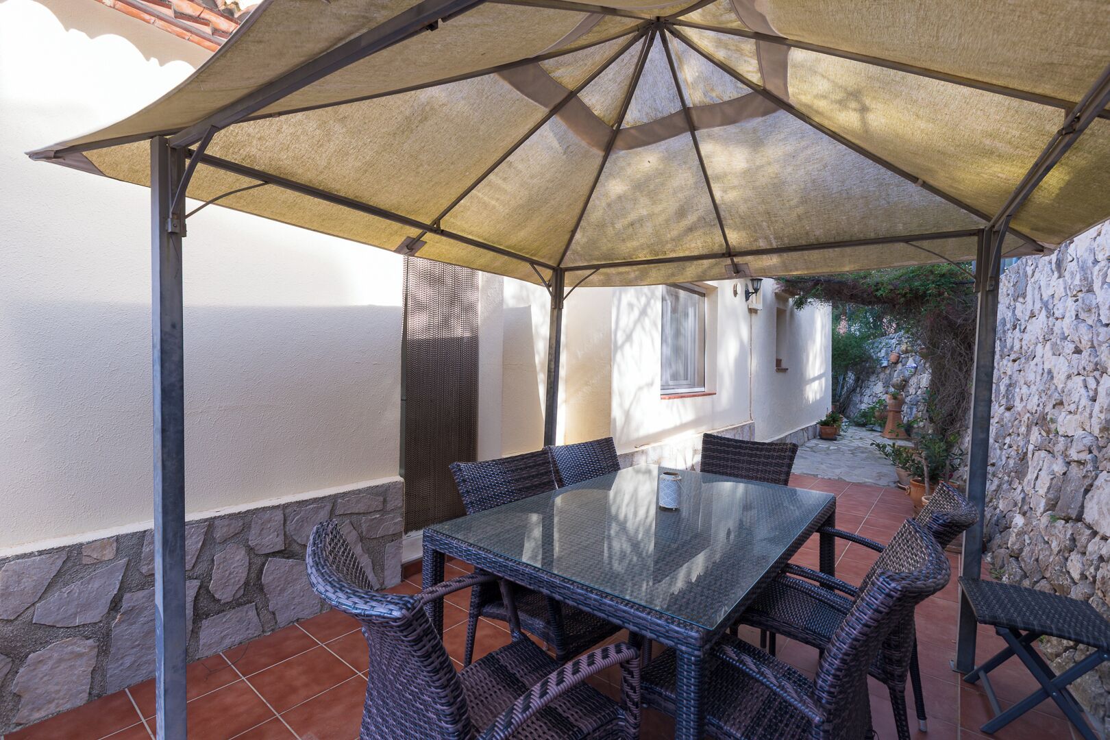 Une terrasse extérieure couverte, dotée d’une table en verre et de chaises en osier, de murs en pierre et offrant une vue sur la mer.