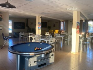 A pool table with balls, surrounded by white chairs and tables in a room with tiled floors and ceiling lights. - Mobile home, pets welcome - 5 guests (Saint-Georges-de-Luzençon)