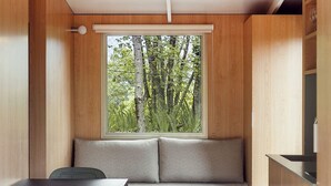 A room with a wooden wall, a window with a view of trees, a sofa, a table, and a chair. - Cozy mobile home, pets welcome - 2 guests (Boltaña)