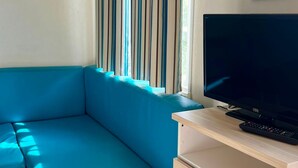 Ein Zimmer mit einem blauen Sofa, einem Fernseher auf einem Holzständer und einem Fenster mit vertikalen Jalousien.