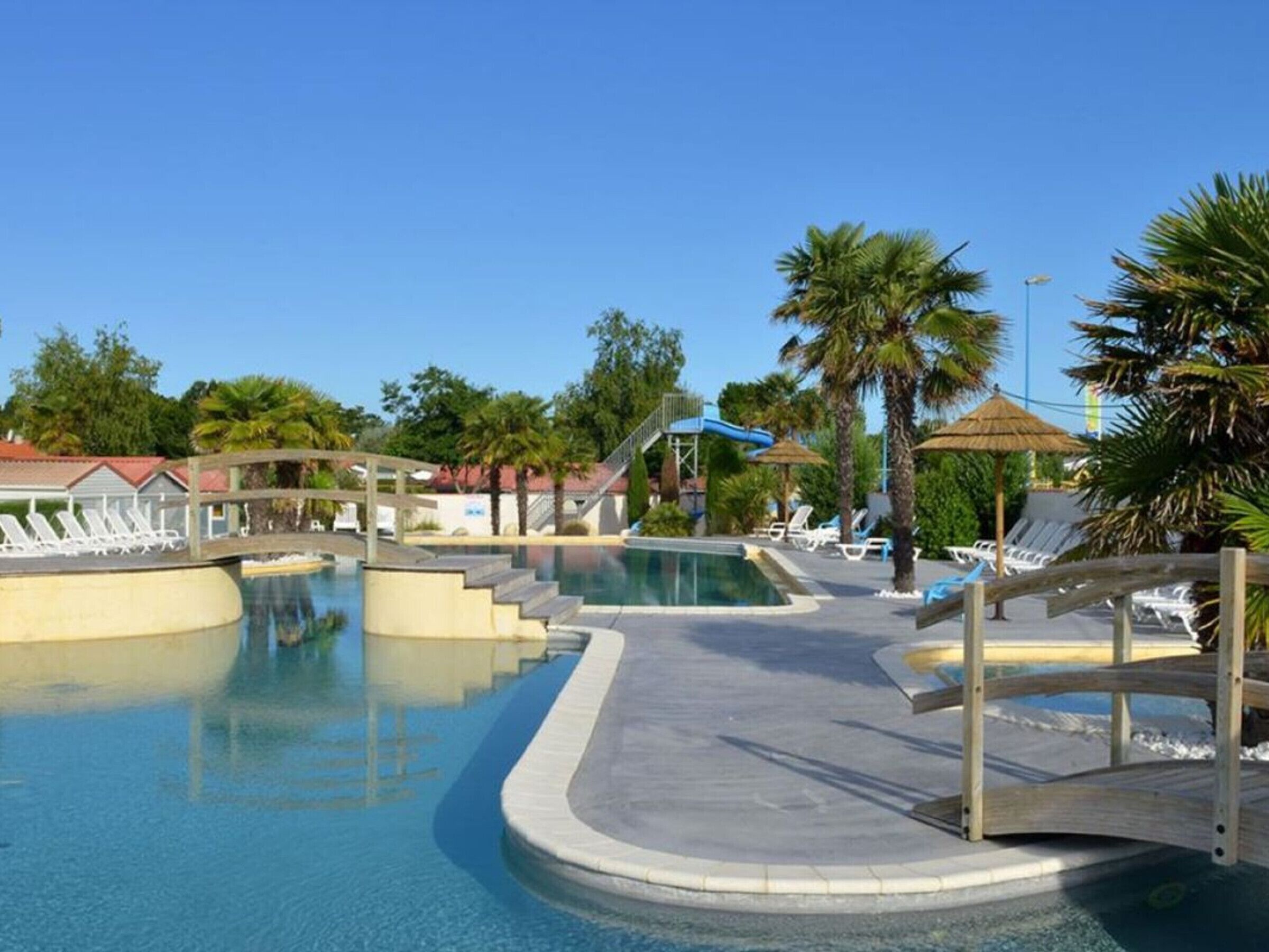 A swimming pool with a wooden bridge, lounge chairs, palm trees, and a clear sky.