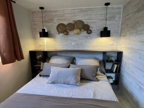 A bedroom with a bed, two bedside tables, a wall-mounted shelf, and a decorative wall piece. - Comfy mobile home - 4 guests (Sarzeau)