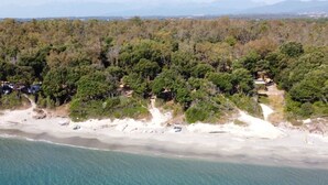 Vista aérea de una zona costera con playa, bosque frondoso y edificios dispersos.
