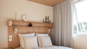 Un dormitorio con cama, mesitas de noche, una mesilla, un ventanal con cortinas y un cabecero de madera con luces fijadas a la pared.