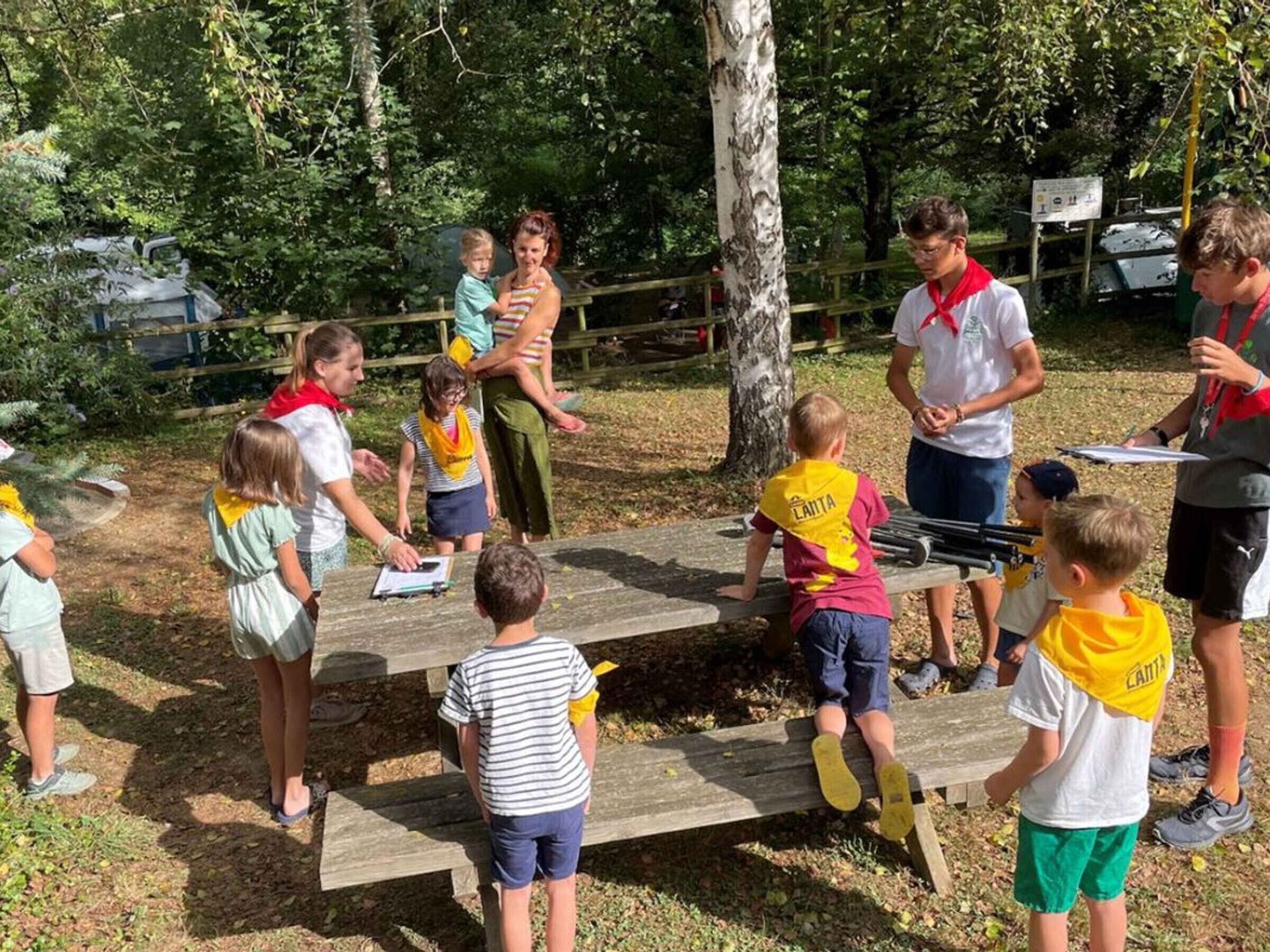 A group of children and adults gathered around wooden benches in an outdoor setting with trees and a fence in the background.