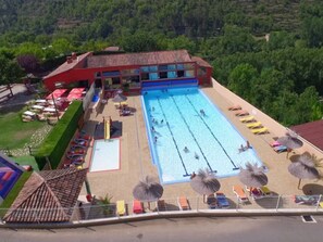 Una piscina con varias pistas, rodeada de tumbonas y sombrillas, con un edificio al fondo.