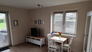 Een kamer met een houten vloer, een witte tafel met stoelen, een televisie op een standaard en een raam met jaloezieën.