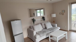 Een kamer met een witte bank, een witte tafel, een witte koelkast, een spiegel, een lamp en een raam met zonwering.